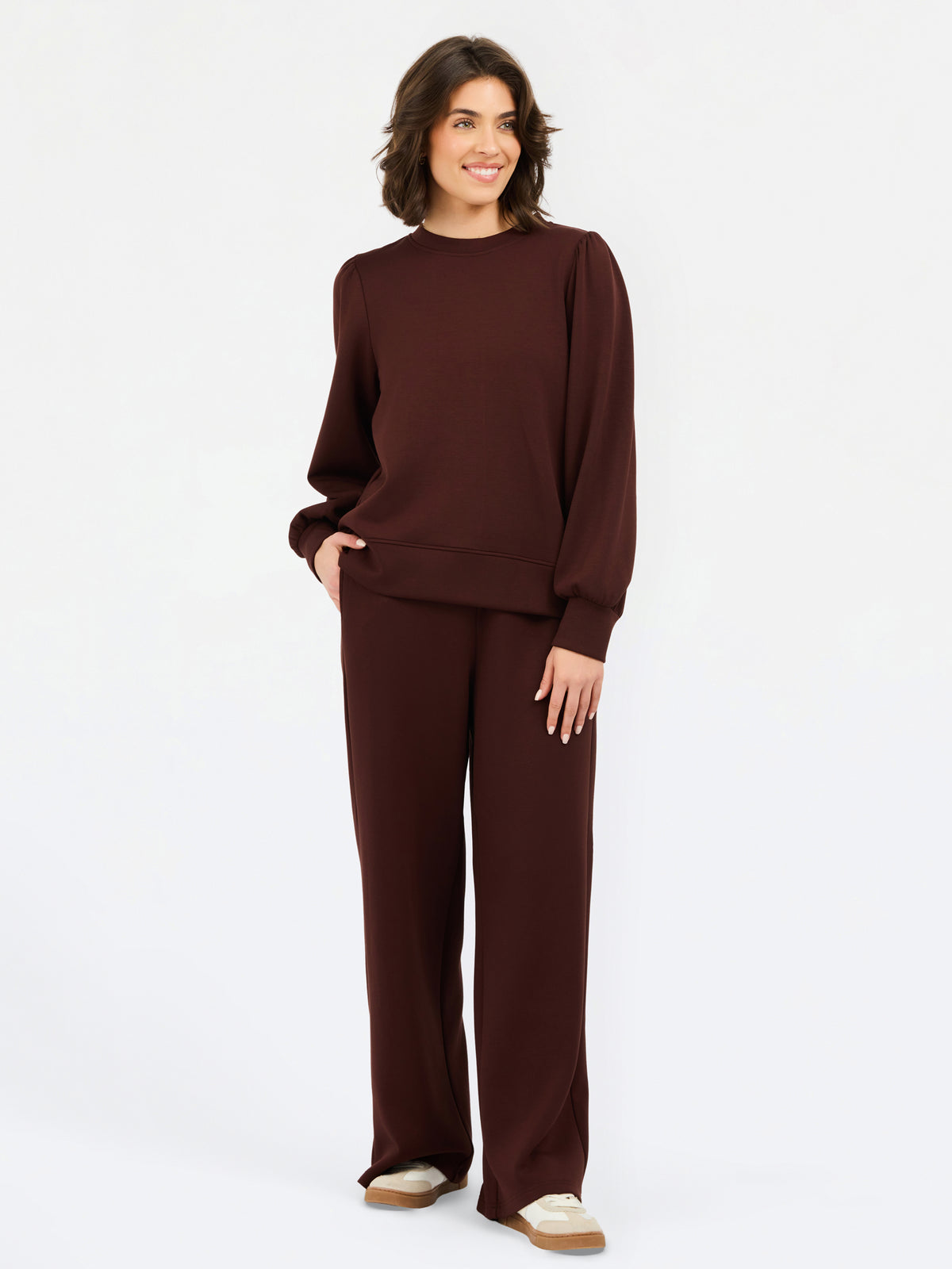 A woman modeling a matching dark brown knit lounge set consisting of a crewneck sweatshirt and wide-leg pants, paired with beige slip-on shoes, against a plain white studio background.