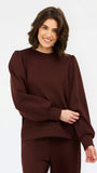 A woman wearing a dark brown crewneck sweatshirt with voluminous balloon sleeves and matching brown pants, photographed against a clean white studio background.
