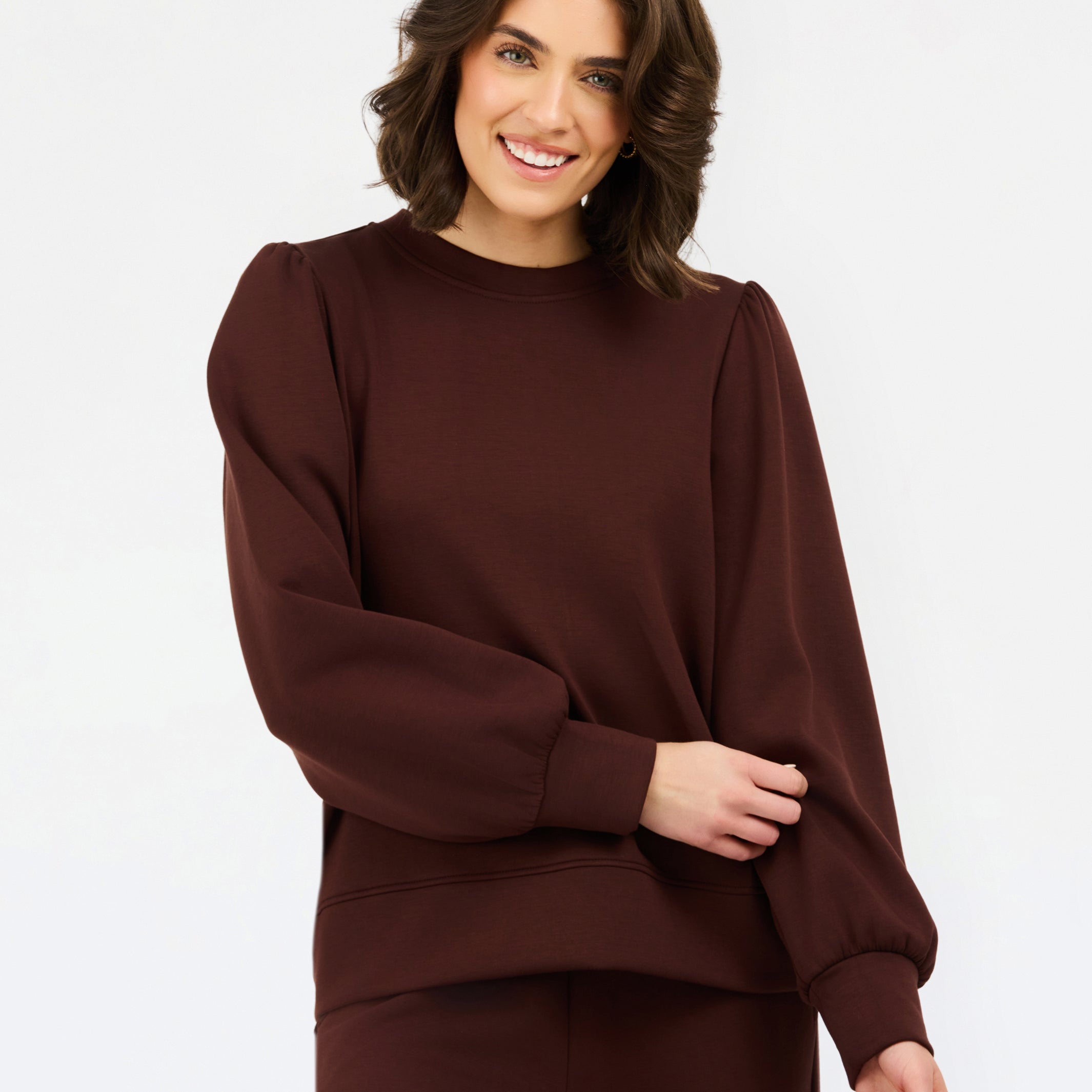 A woman wearing a dark brown crewneck sweatshirt with voluminous balloon sleeves and matching brown pants, photographed against a clean white studio background.