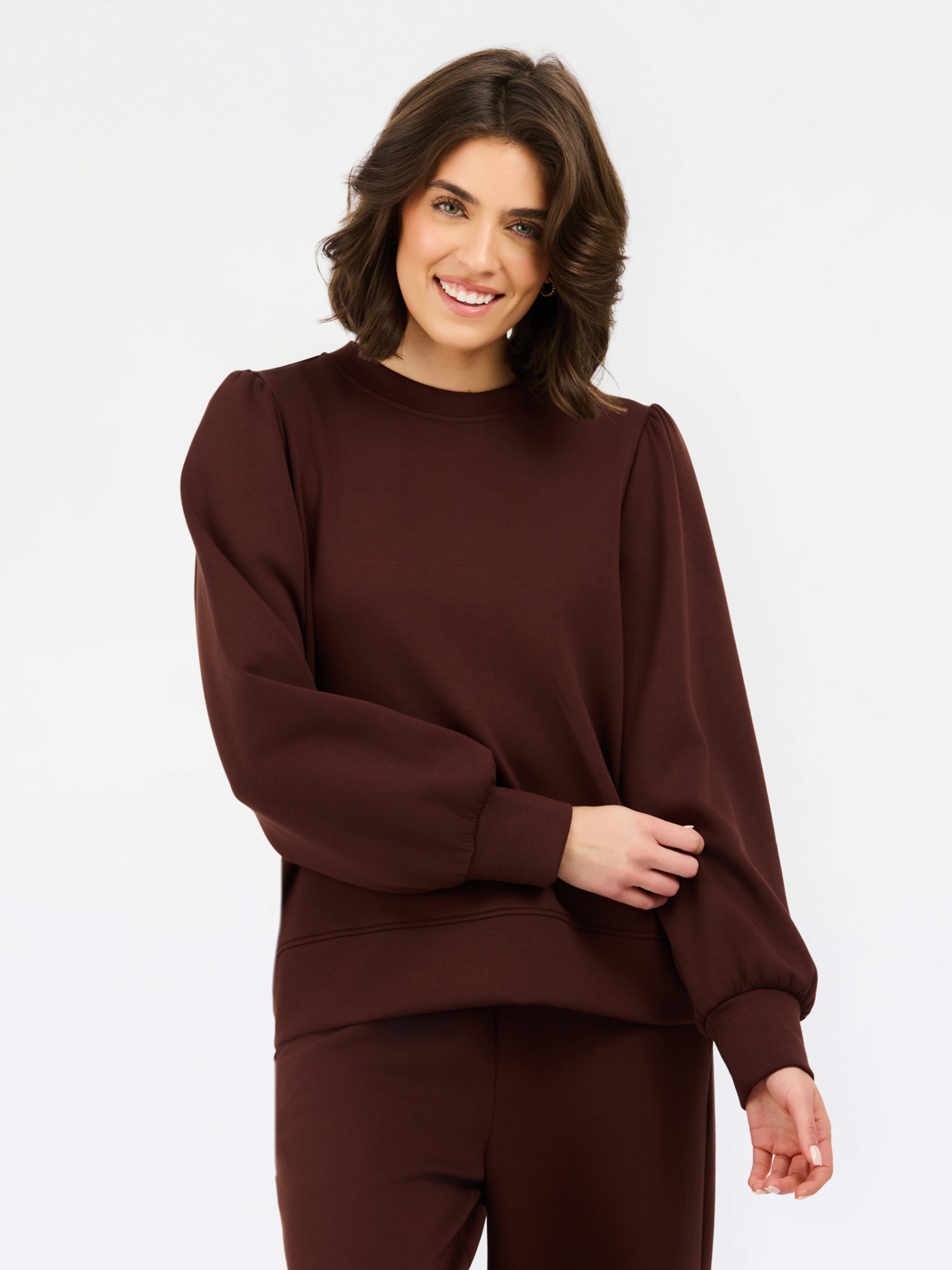 A woman wearing a dark brown crewneck sweatshirt with voluminous balloon sleeves and matching brown pants, photographed against a clean white studio background.