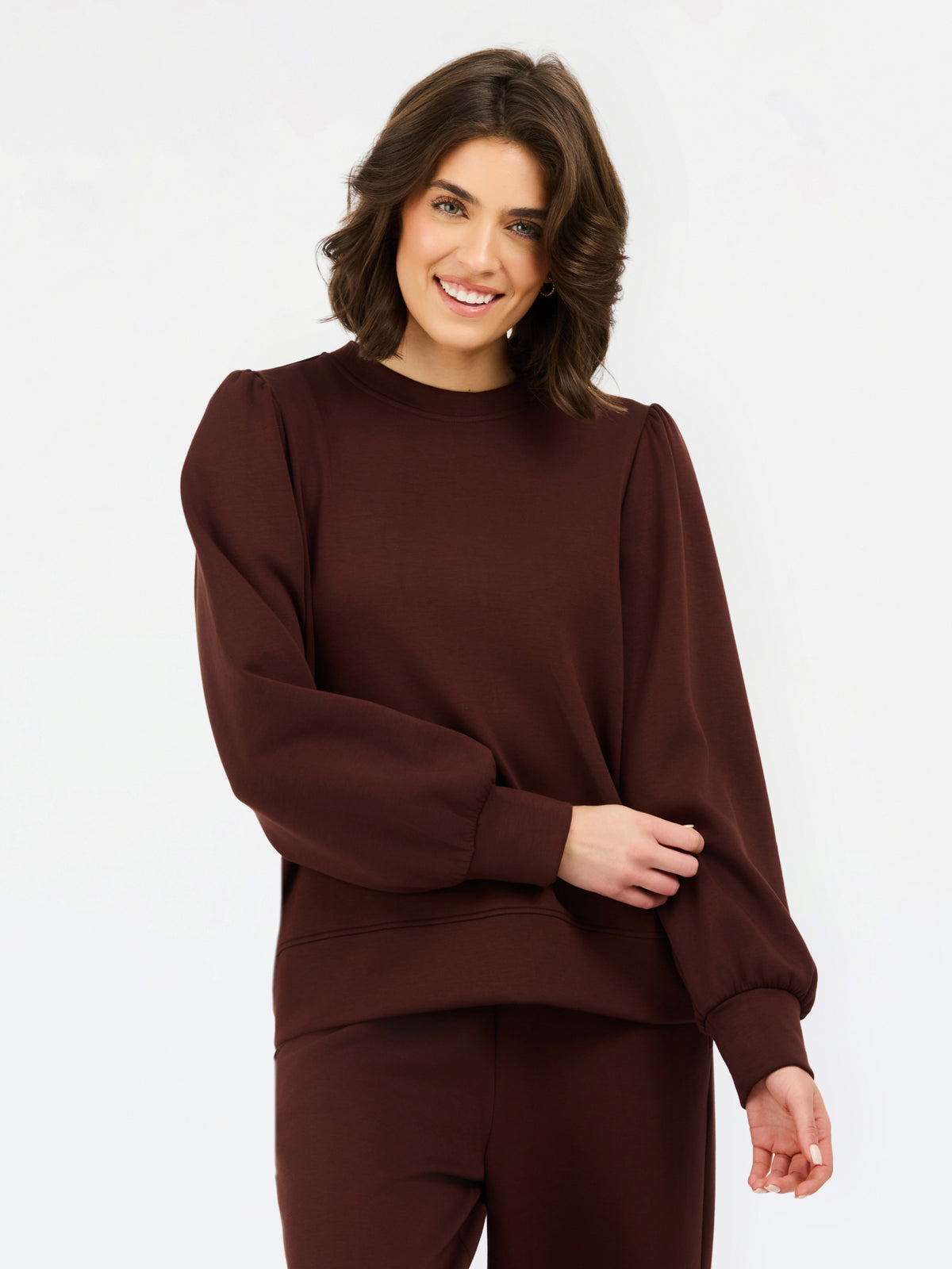 A woman wearing a dark brown crewneck sweatshirt with voluminous balloon sleeves and matching brown pants, photographed against a clean white studio background.
