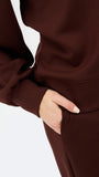 Close-up of a person wearing a dark brown long-sleeve top or dress, with one hand tucked into a side pocket against a plain light background.