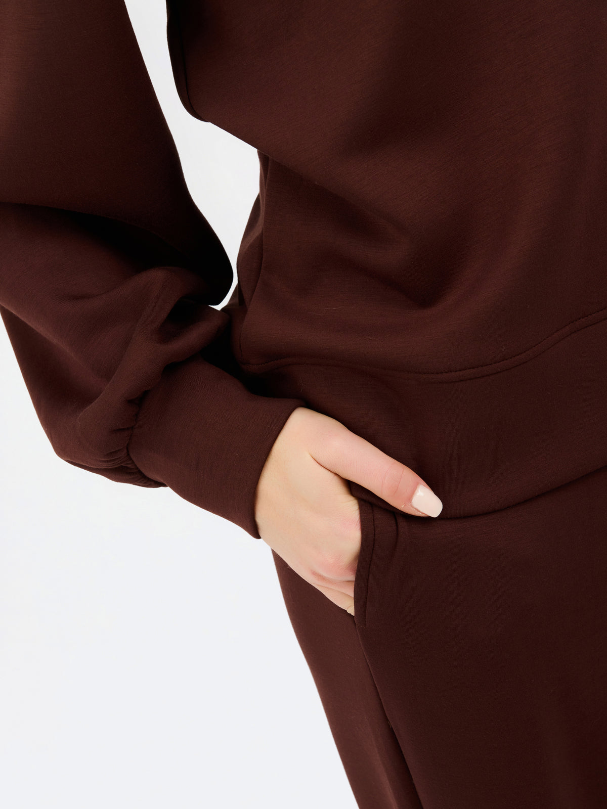 Close-up of a person wearing a dark brown long-sleeve top or dress, with one hand tucked into a side pocket against a plain light background.