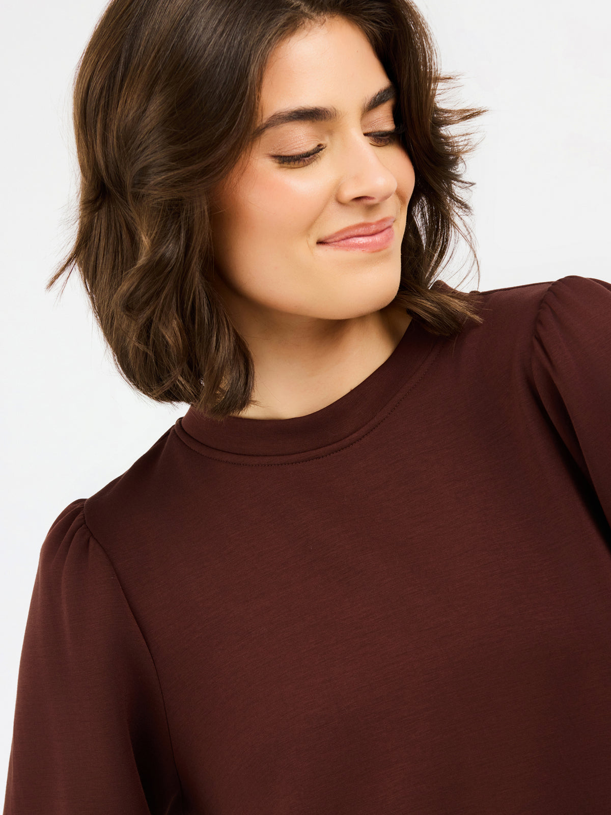 A close-up shot of a woman wearing a dark brown long-sleeve top with puffed shoulders, set against a plain light gray background.