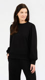 A woman modeling a black crewneck sweatshirt with balloon sleeves, paired with black pants, against a plain white studio background.