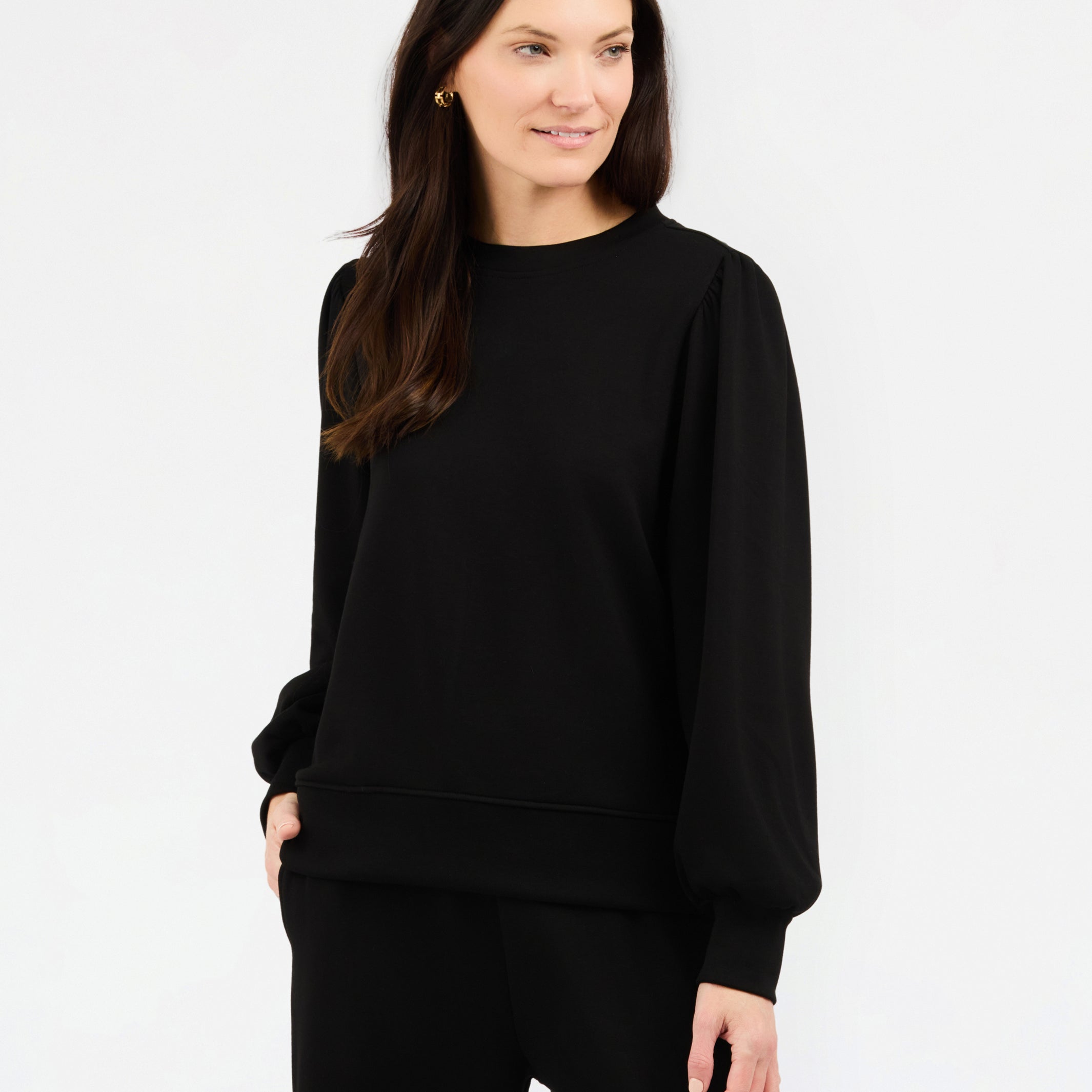 A woman modeling a black crewneck sweatshirt with balloon sleeves, paired with black pants, against a plain white studio background.