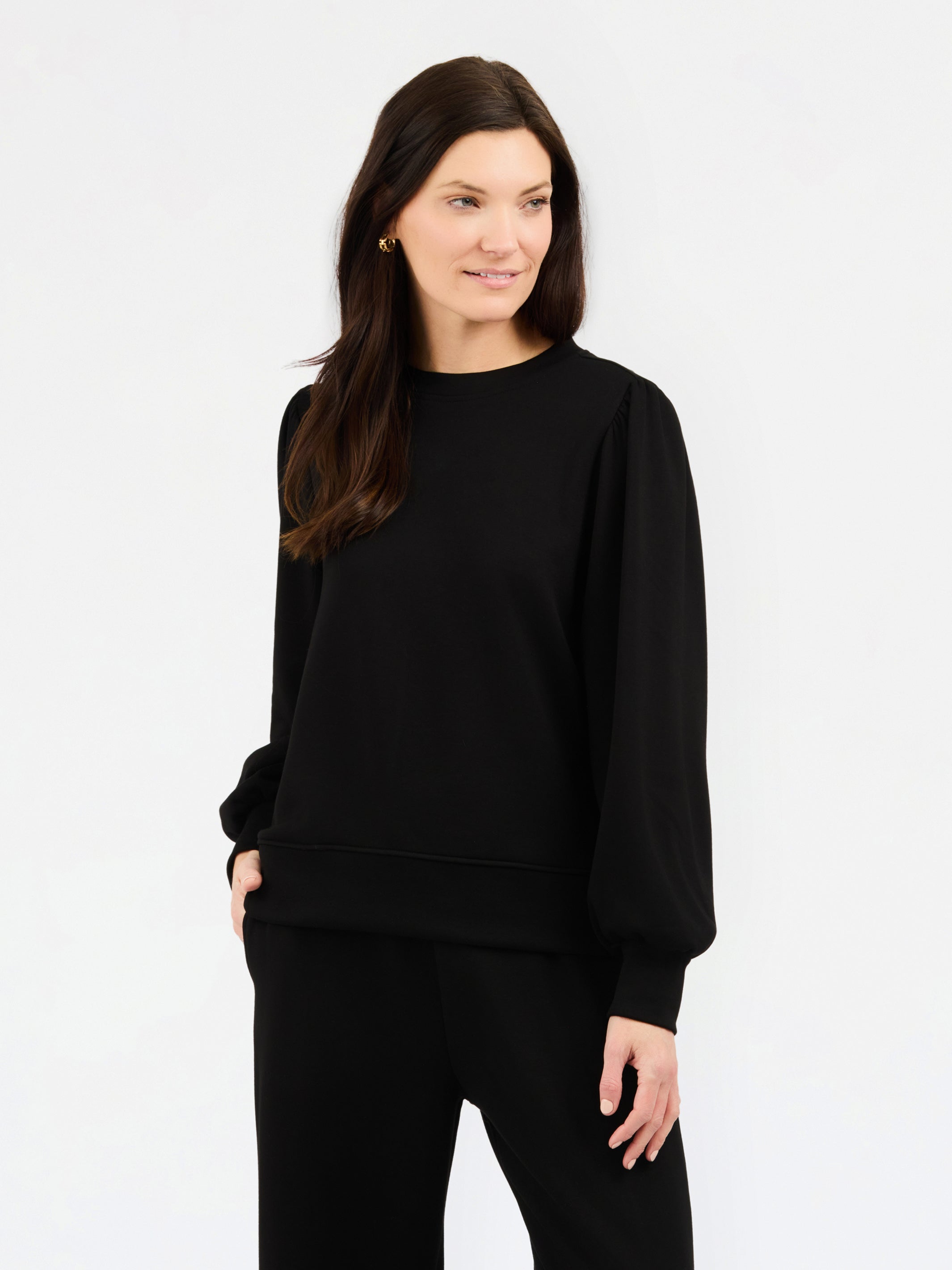 A woman modeling a black crewneck sweatshirt with balloon sleeves, paired with black pants, against a plain white studio background.