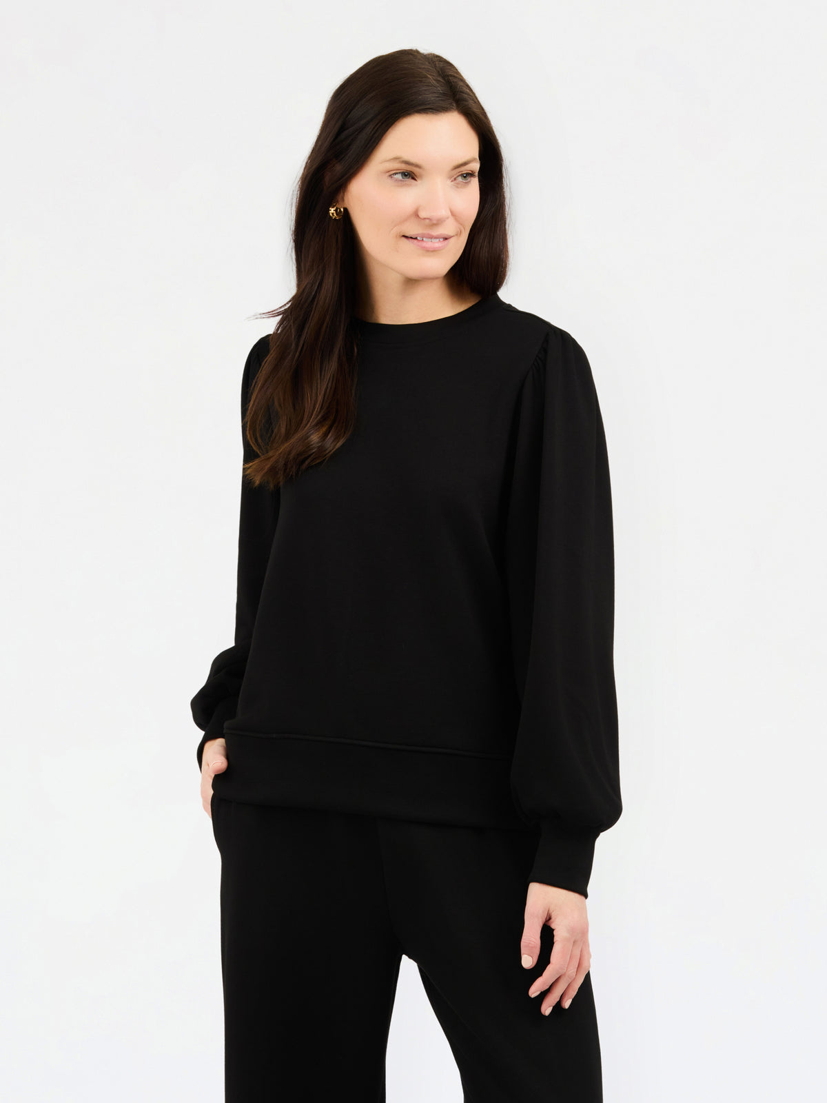 A woman modeling a black crewneck sweatshirt with balloon sleeves, paired with black pants, against a plain white studio background.