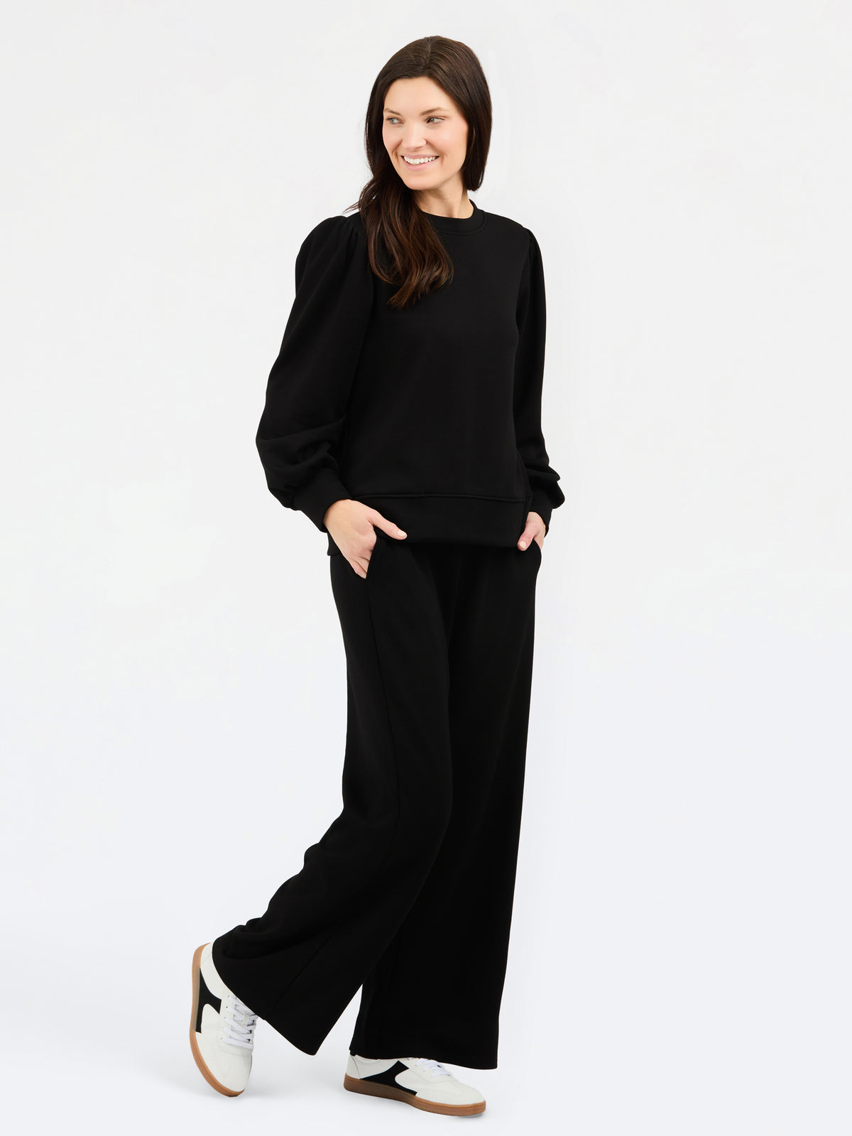 A woman modeling a black long-sleeve sweatshirt with balloon sleeves and matching black wide-leg pants, paired with white sneakers, against a plain white studio background.