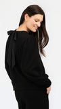 A woman wearing a black long-sleeve top with a large bow detail on the back, shown from a side-back angle against a plain white studio background.