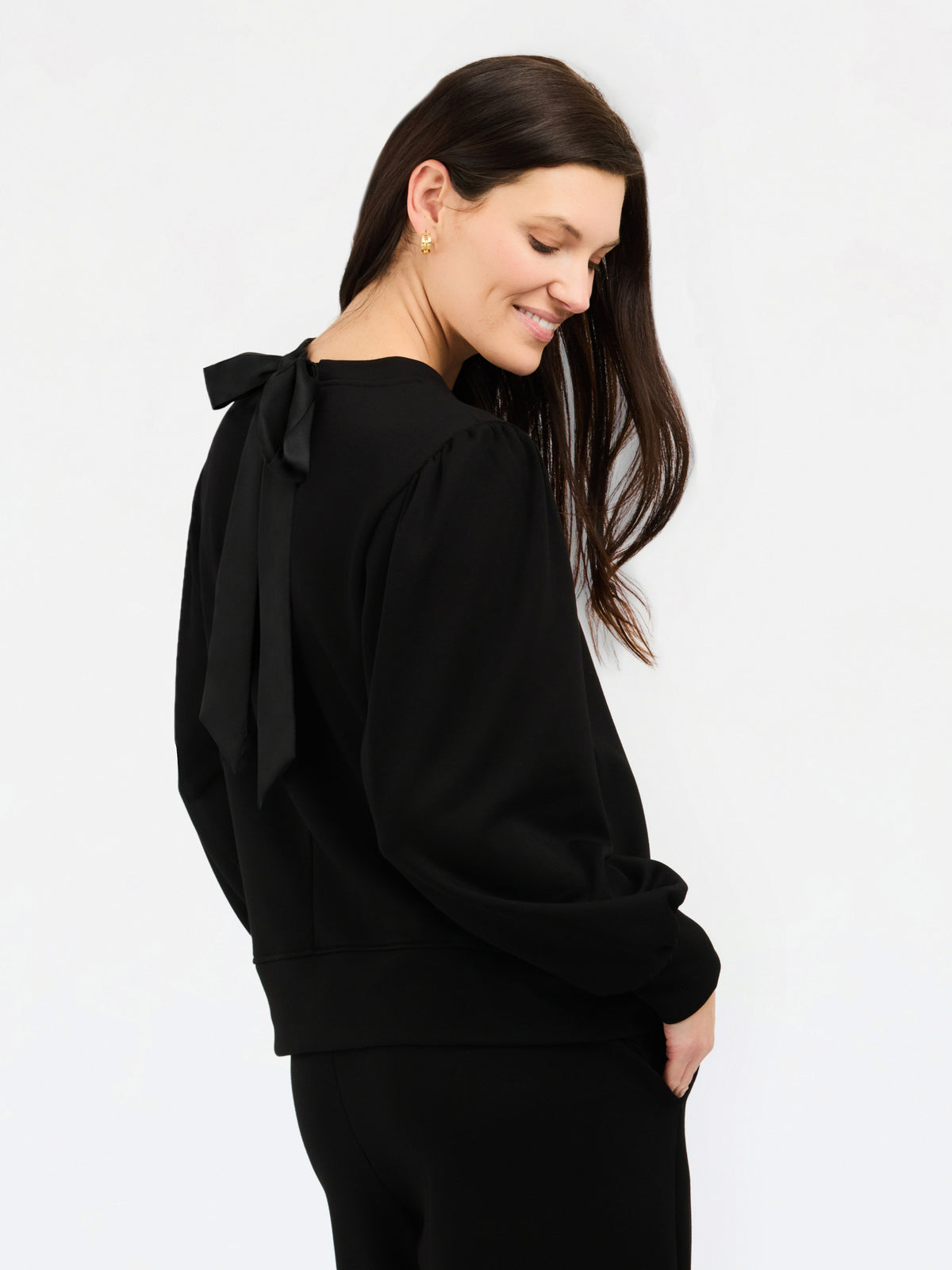 A woman wearing a black long-sleeve top with a large bow detail on the back, shown from a side-back angle against a plain white studio background.