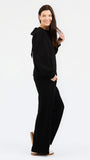 A woman modeling a black long-sleeve set with a wide collar and draped shoulder detail, paired with tan sneakers against a plain white studio background.