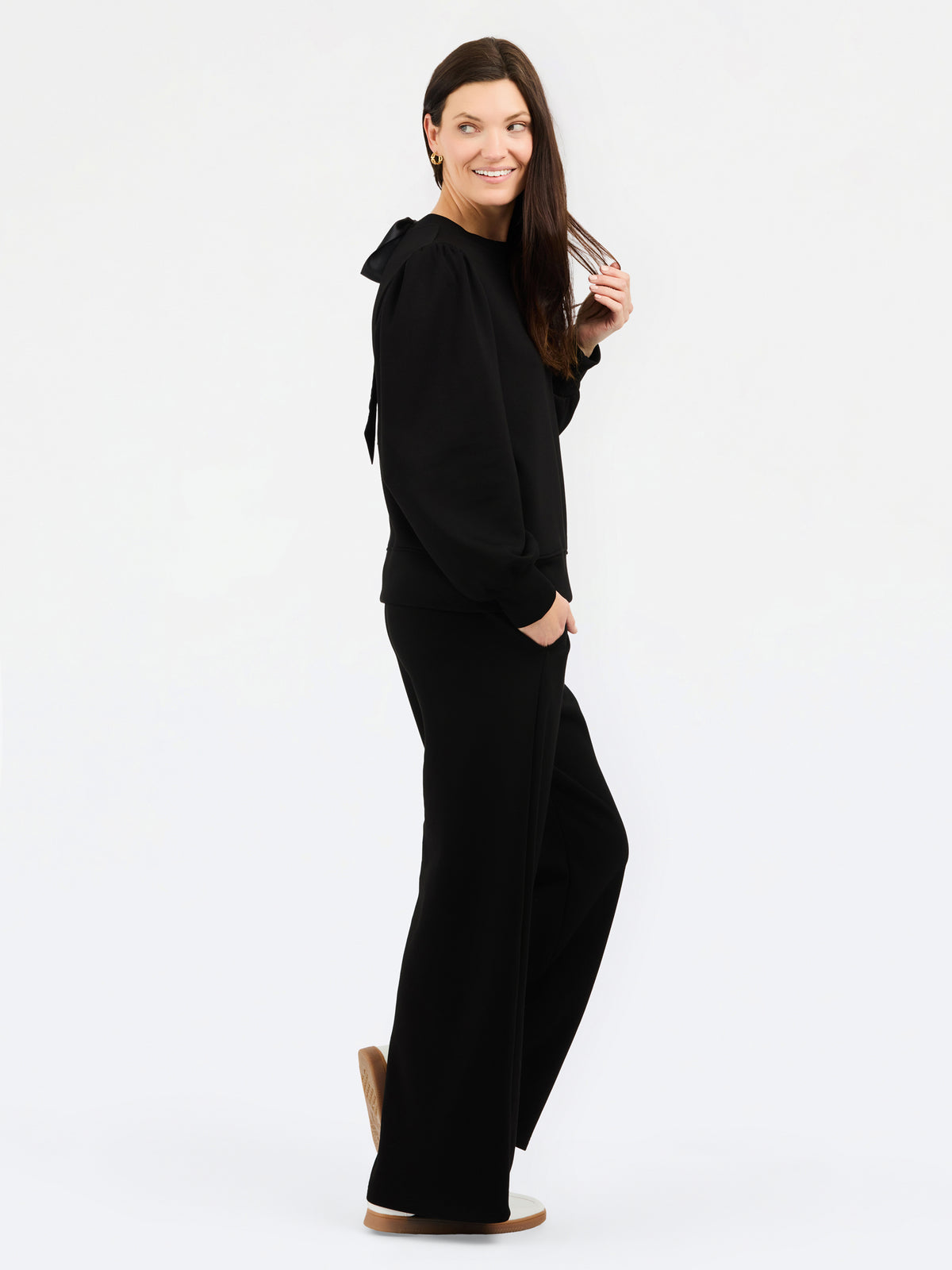 A woman modeling a black long-sleeve set with a wide collar and draped shoulder detail, paired with tan sneakers against a plain white studio background.