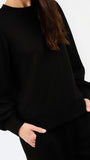 A close-up of a person wearing a black crewneck sweatshirt with voluminous long sleeves and matching black pants, with hands in pockets, against a plain white background.
