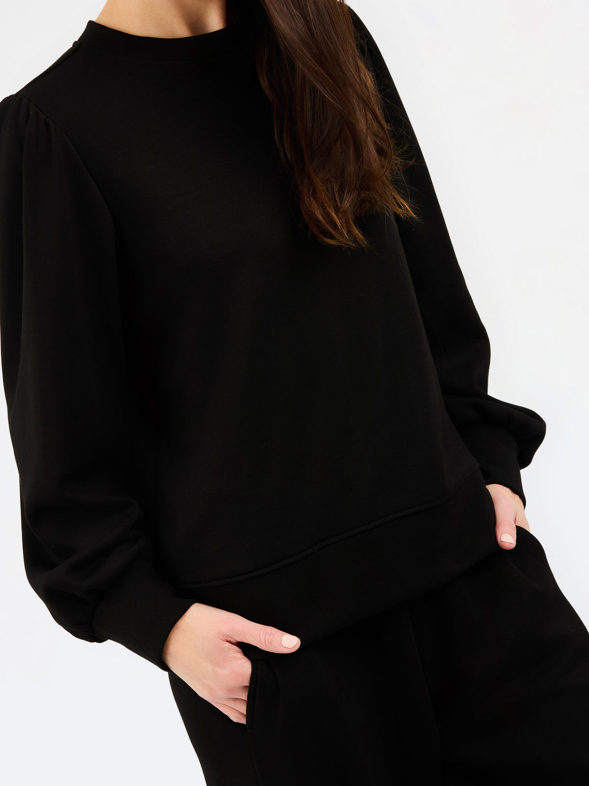A close-up of a person wearing a black crewneck sweatshirt with voluminous long sleeves and matching black pants, with hands in pockets, against a plain white background.