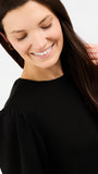 A close-up of a smiling woman with long dark brown hair wearing a black long-sleeve top with gathered shoulders, set against a plain light gray background.