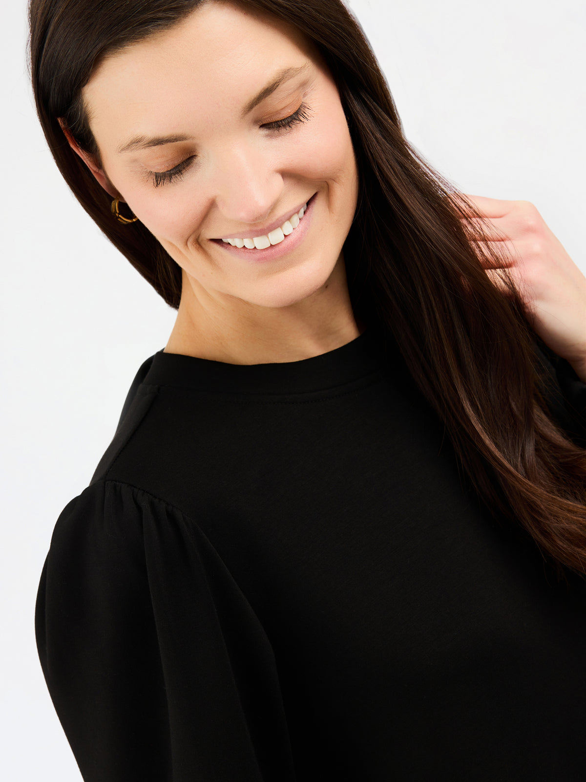 A close-up of a smiling woman with long dark brown hair wearing a black long-sleeve top with gathered shoulders, set against a plain light gray background.