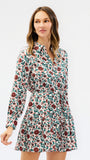 A woman modeling a long-sleeve mini dress with a dense floral print in red, green, and white, set against a plain light gray studio background.