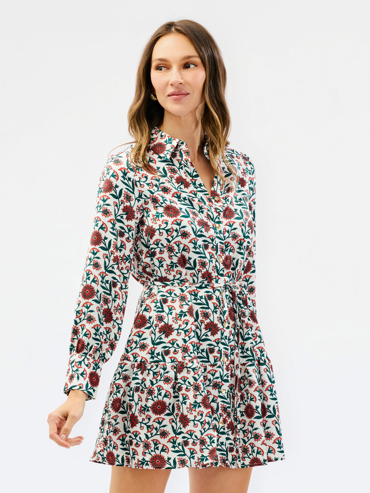 A woman modeling a long-sleeve mini dress with a dense floral print in red, green, and white, set against a plain light gray studio background.
