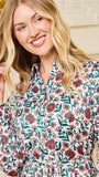 A smiling blonde woman wearing a long-sleeve button-up blouse with a dense red, green, and white floral pattern, posed against a soft beige background.