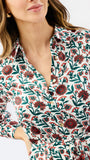 A close-up of a woman wearing a white button-up shirt with a red and green floral print. The image is cropped to show her from the mouth down to the waist, with her hand in her pocket.