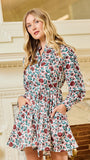 A woman modeling a long-sleeve floral mini dress with pockets, standing in front of an ornate cream-colored fireplace.