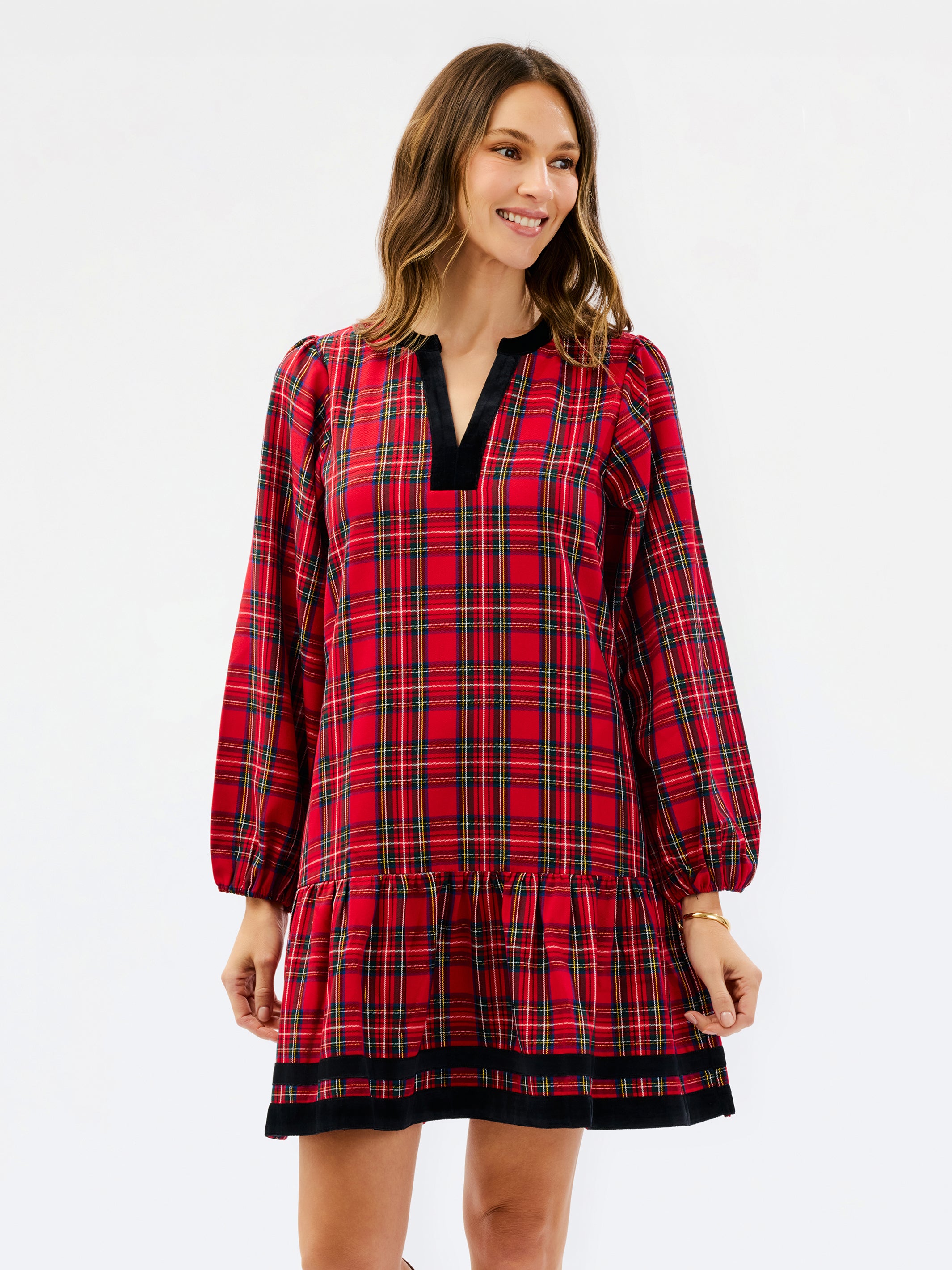 A woman modeling a red tartan plaid mini dress with black velvet trim at the split neckline and hem, set against a plain white studio background.