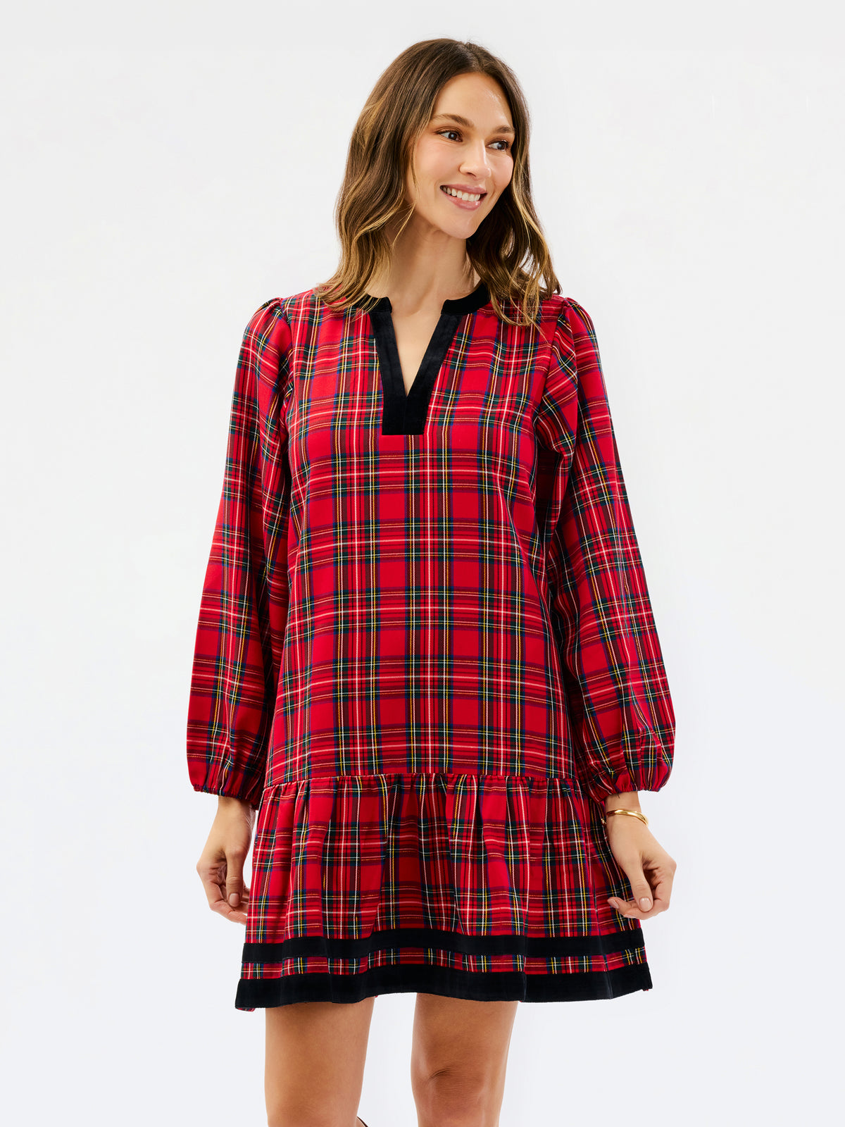 A woman modeling a red tartan plaid mini dress with black velvet trim at the split neckline and hem, set against a plain white studio background.