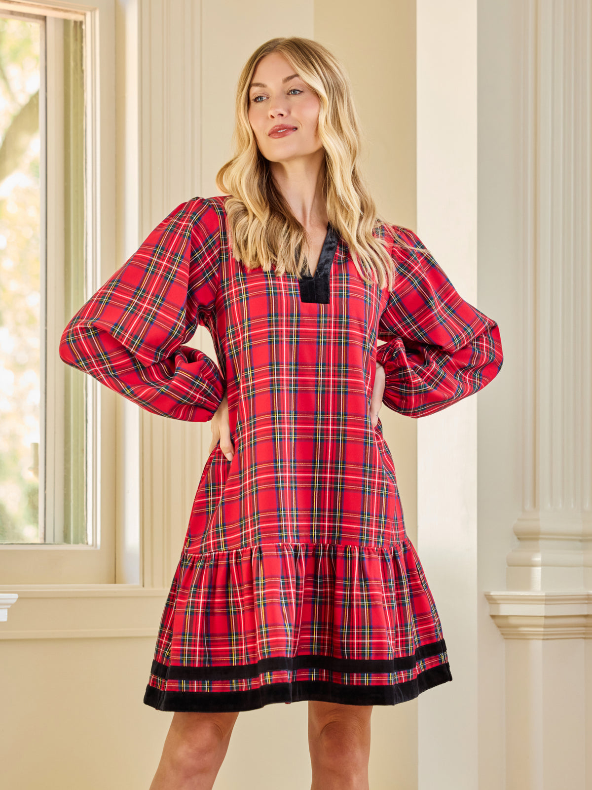 A woman modeling a red tartan plaid nightgown with black trim, standing in a brightly lit room with cream-colored columns and a window.