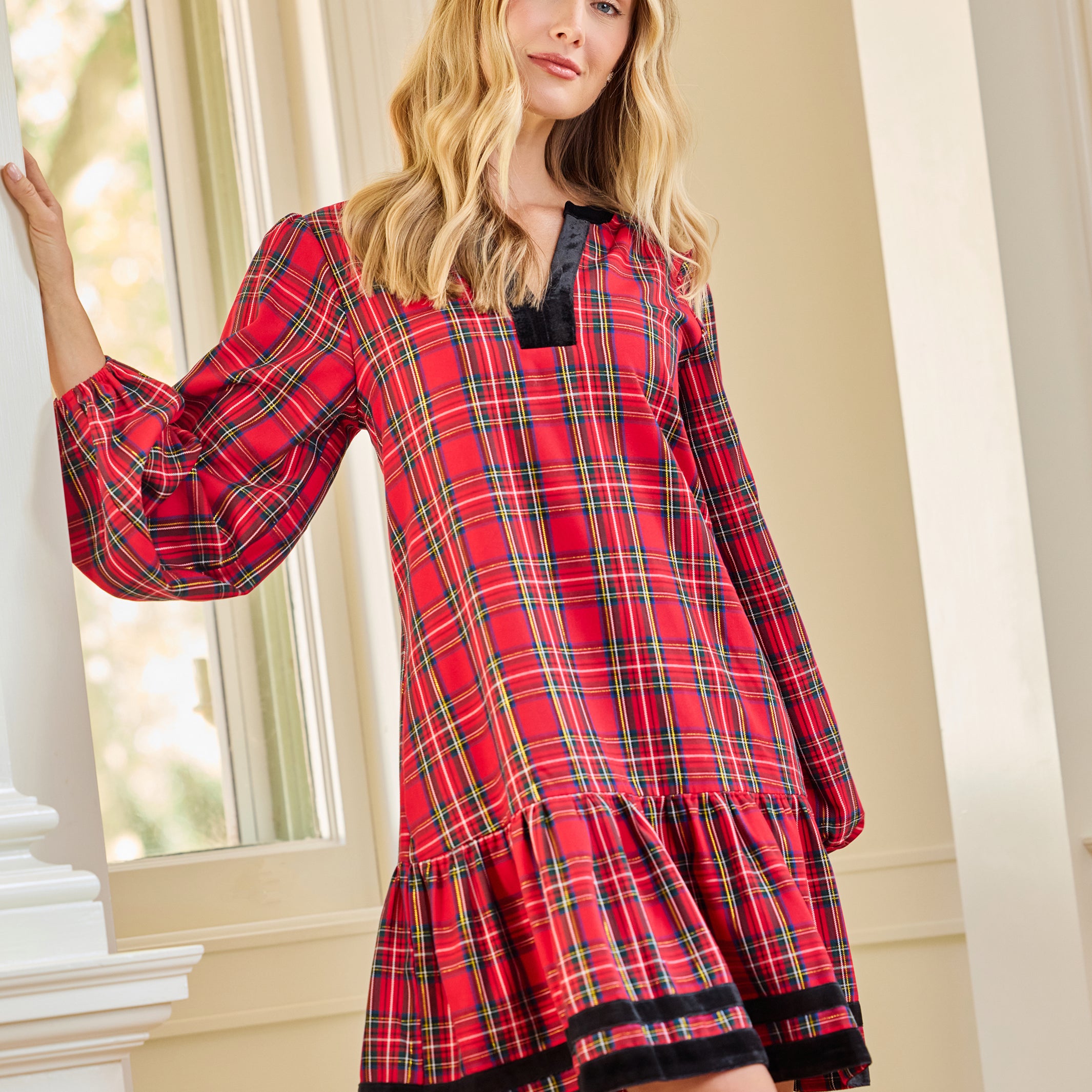 A woman wearing a red and black plaid long-sleeve nightgown with a tiered hem, posing indoors near a window.