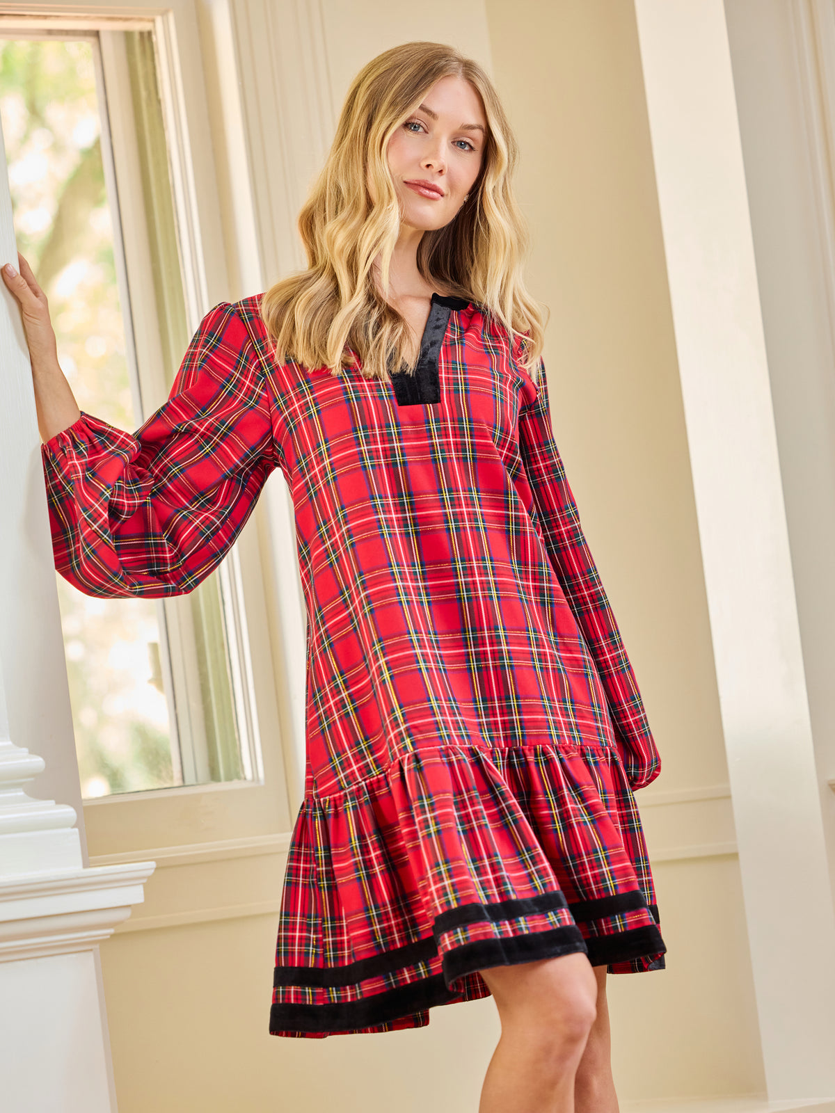 A woman wearing a red and black plaid long-sleeve nightgown with a tiered hem, posing indoors near a window.