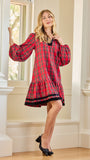 A woman modeling a red and black plaid long-sleeve tunic dress with a ruffled hem, standing in a bright room with white columns and a window.
