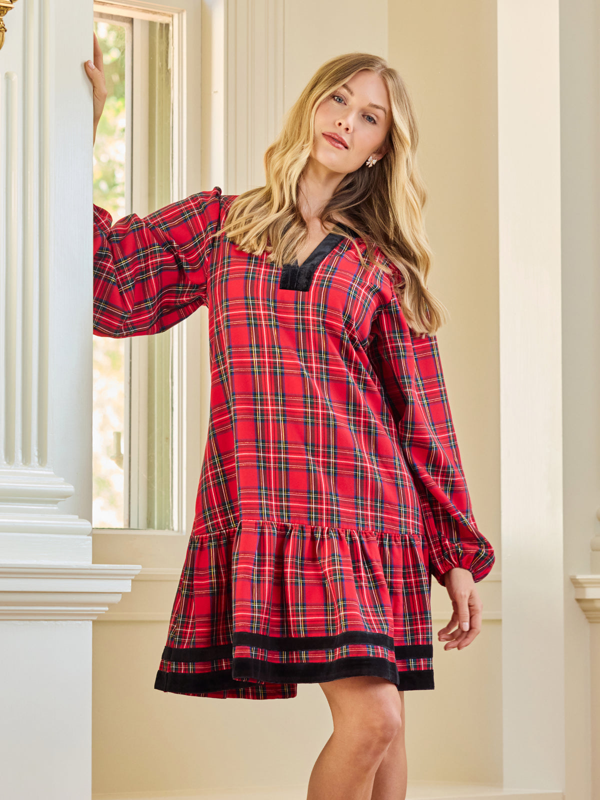 A woman wearing a red and black plaid long-sleeve nightgown or tunic dress, posing in front of a white classical column and window.