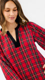 A woman wearing a red plaid long-sleeve top with a black V-neck trim, shown against a plain light gray studio background.