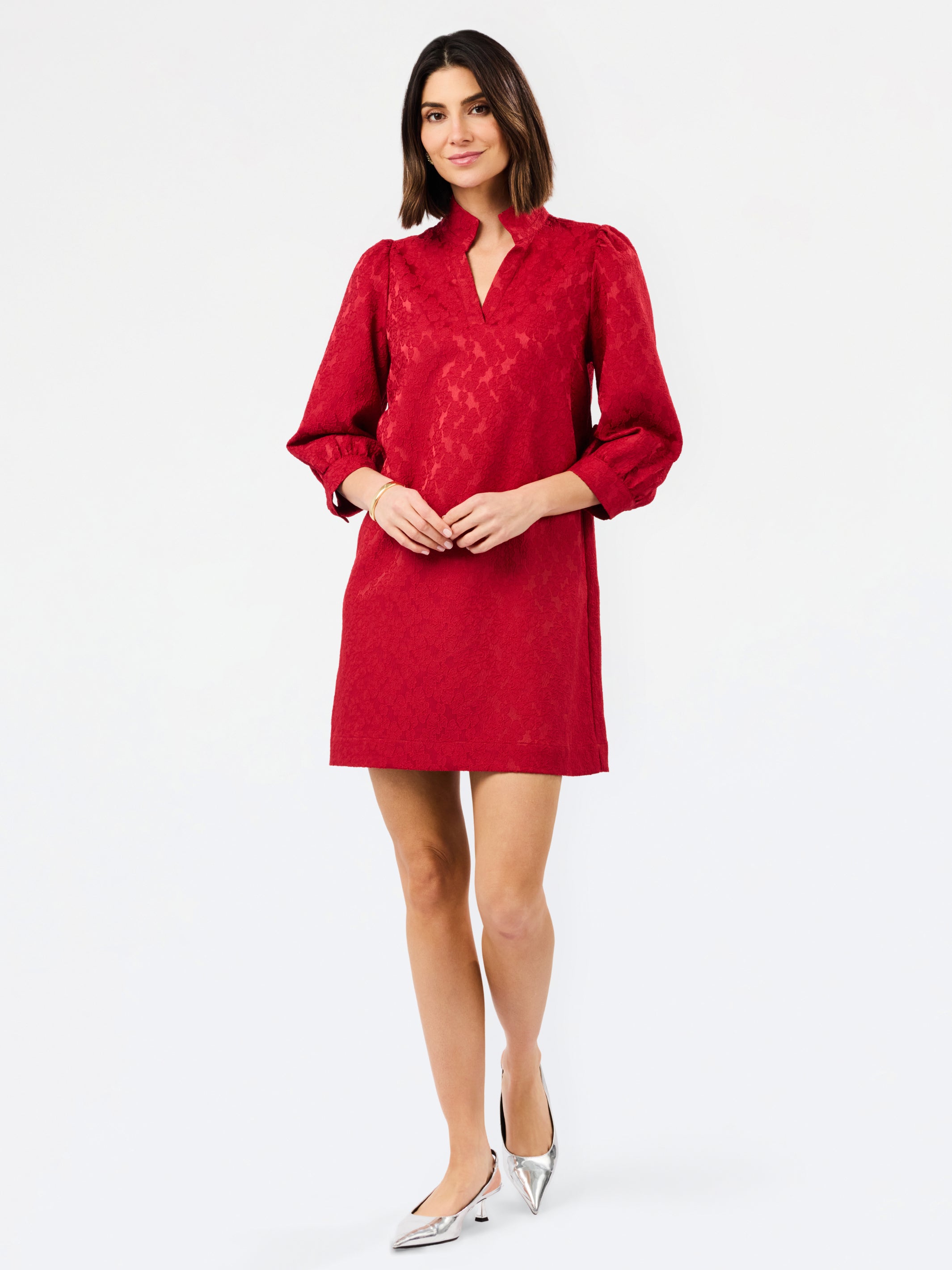 A woman modeling a red textured mini dress with three-quarter sleeves and a split neckline, paired with silver pointed-toe heels, against a plain white studio background.