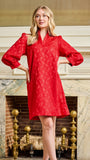 A woman modeling a bright red lace tunic dress with long puff sleeves and a V-neckline, standing in front of an ornate fireplace.