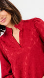 A close-up of a woman wearing a textured red blouse with a high split neckline and long sleeves, photographed against a plain light background.