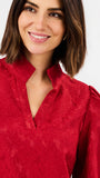 A close-up of a woman wearing a textured red blouse with a high mock neck and puffed shoulders, set against a plain white background.