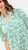 A woman wearing a short-sleeve collared blouse with a green and white geometric pattern, shown in a close-up studio shot against a light gray background.
