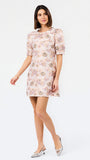 A woman modeling a short pink floral dress with puff sleeves and a ruffled neckline, paired with metallic silver heels, against a plain white studio background.