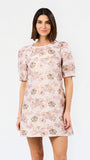 A woman modeling a short-sleeve shift dress with a light pink base and a multicolor floral pattern, set against a plain white studio background.