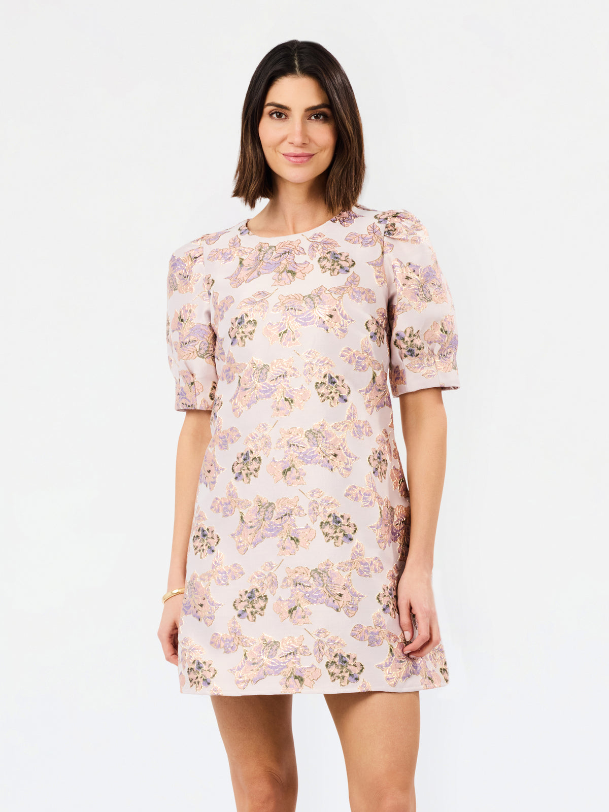 A woman modeling a short-sleeve shift dress with a light pink base and a multicolor floral pattern, set against a plain white studio background.