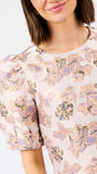 A close-up of a woman wearing a short-sleeve blouse with a soft floral print in pastel pink, lavender, and green on a light cream background.