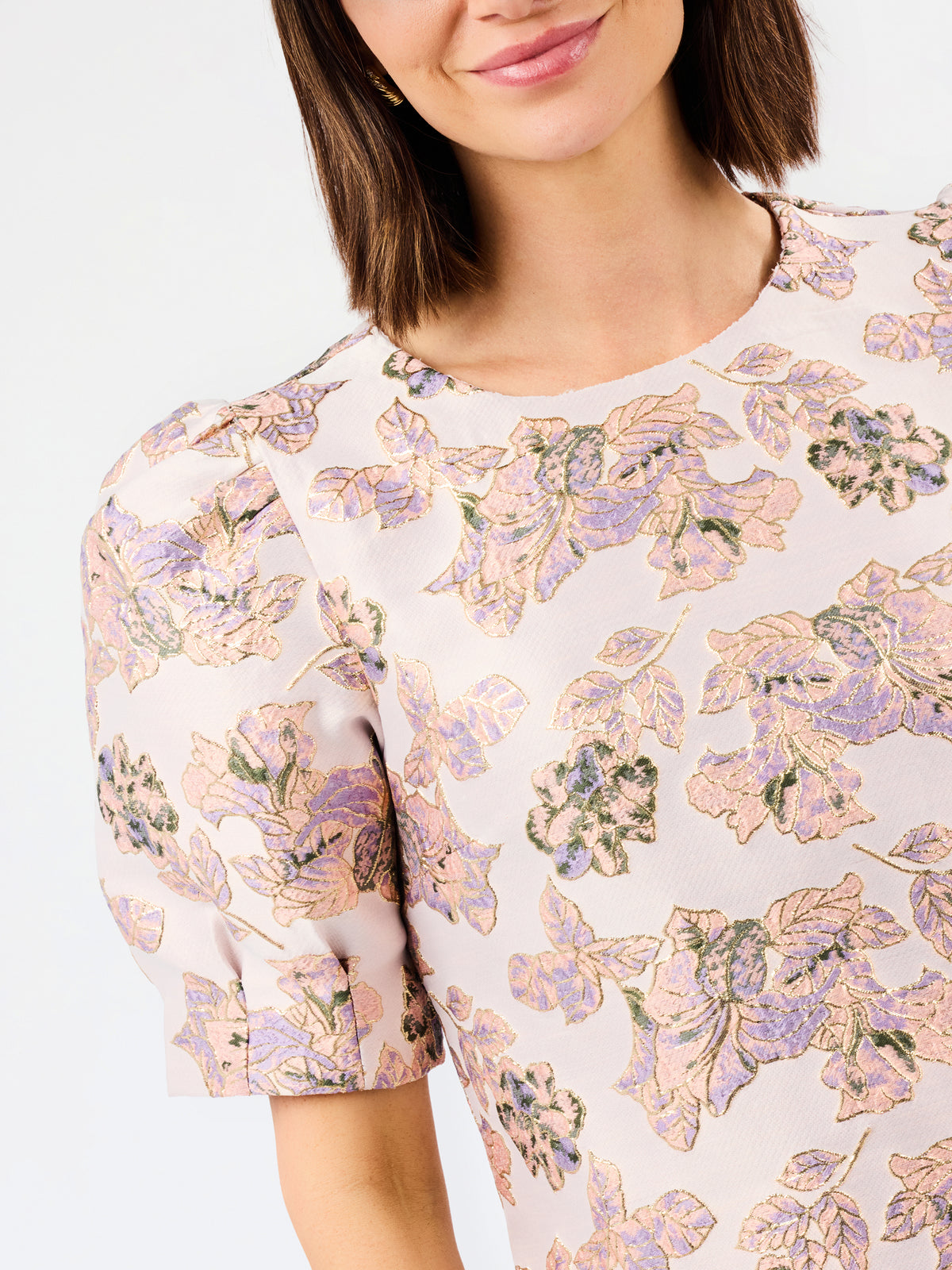 A close-up of a woman wearing a short-sleeve blouse with a soft floral print in pastel pink, lavender, and green on a light cream background.