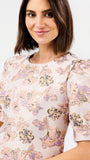 A woman wearing a short-sleeve blouse with a soft floral pattern in pastel pink, lavender, and beige tones, photographed against a plain light background.