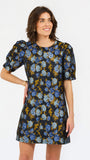 A woman modeling a short-sleeve A-line dress with a blue and mustard yellow floral print on a black background, set against a plain white studio background.