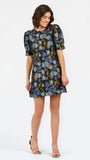 A woman modeling a short puff-sleeve mini dress with a blue, yellow, and white floral pattern on a dark background, paired with beige strappy heels, against a plain white studio background.