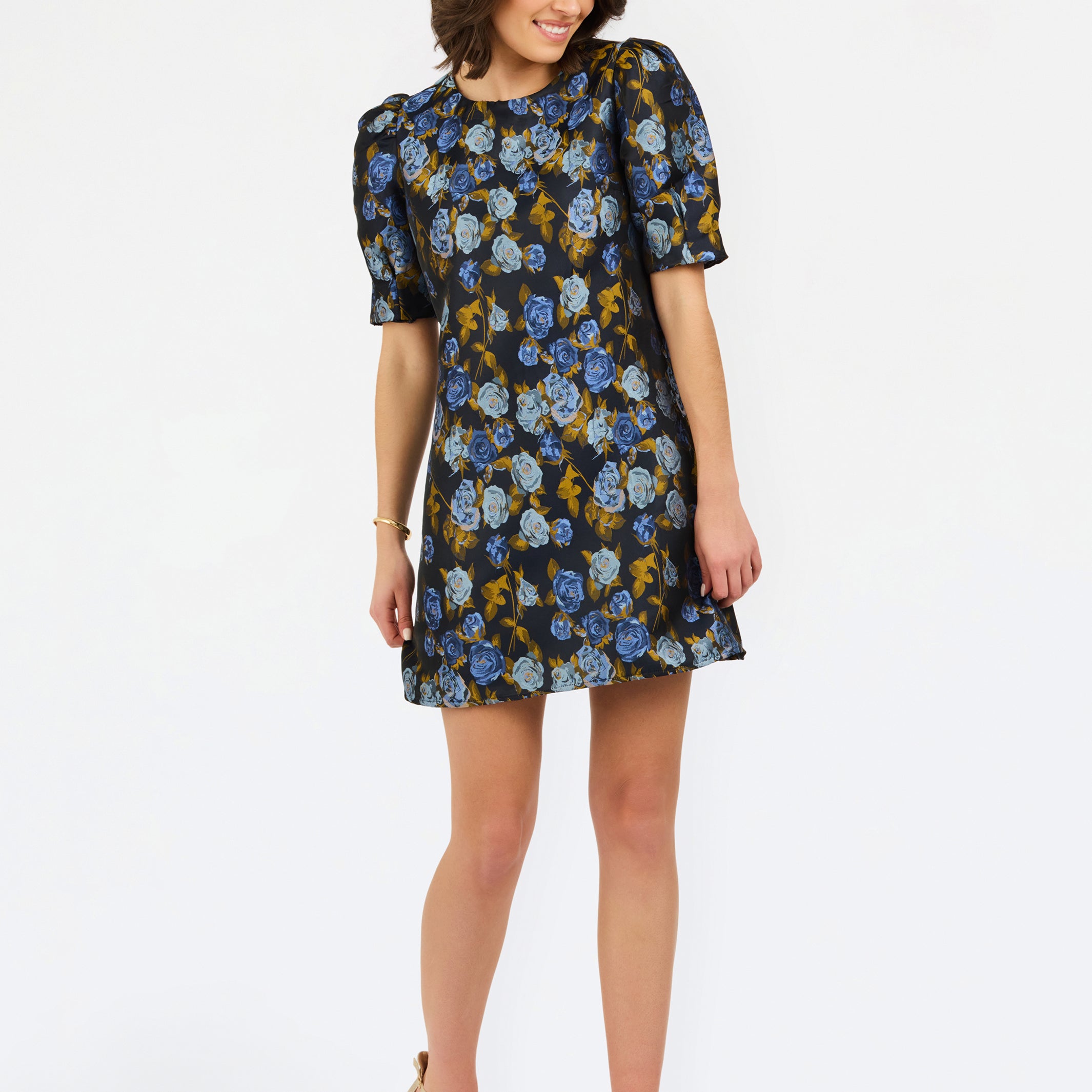 A woman modeling a short puff-sleeve mini dress with a blue, yellow, and white floral pattern on a dark background, paired with beige strappy heels, against a plain white studio background.