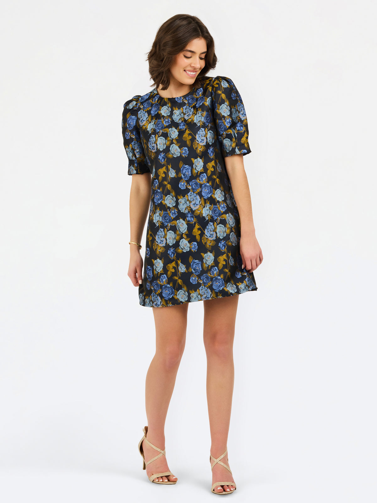 A woman modeling a short puff-sleeve mini dress with a blue, yellow, and white floral pattern on a dark background, paired with beige strappy heels, against a plain white studio background.