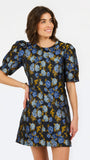 A woman modeling a short puff-sleeve dress with an all-over floral print in blue, light blue, and mustard yellow on a dark background, set against a plain white studio backdrop.